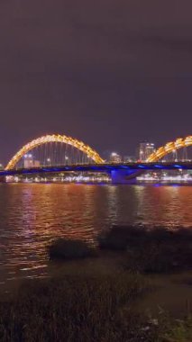 Dikey video. Altın Ejder Köprüsü Yansıtıcı Nehir 'in üzerinde parlıyor. Şehir Geniş Açı Görüş Alanı' nda, Renkli Gece Işıkları ve Pürüzsüz Hareket, Da Nang Zaman kavramını yakalıyor.