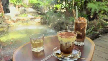 Geniş Açı Yavaş Hareket Görünümü Yumurta Kahvesi ve Buzlu İçecekler Tahta Masa üzerinde Parlak Bahçe Göletinin yanında Düşen Su Rahatlatıcı bir Kafe Konsepti oluşturur