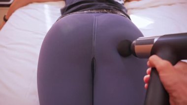 Erkek, kadının üzerine eğilir kalçasına masaj yapar dar taytlı masaj tabancasıyla yavaş çekim titreşim dalgalanması, spor kurtarma konsepti...