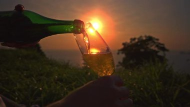 Ellerin Yavaş Çekimi Yeşil Şişe 'den Uzun Cam' e köpüklü şarap dökerken Sıvı ve Yumuşak Sahil Arkaplanı 'na hizalanmış altın günbatımı, Kutlama Konsepti