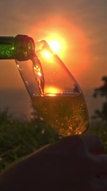 Dikey video. Ellerin Yavaş Çekimi Yeşil Şişe 'den Uzun Cam' e köpüklü şarap dökerken Sıvı ve Yumuşak Sahil Arkaplanı 'na hizalanmış altın günbatımı, Kutlama Konsepti