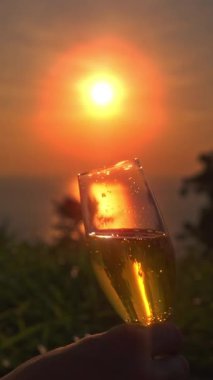 Dikey video. Yavaş Çekimde Yavaş Çekim Şampanya Kadehi ile Parlayan Portakal Günbatımına doğru Sakin Deniz 'in üzerinde Yumuşak Çimen Önplanı, Boş Zaman Konsepti