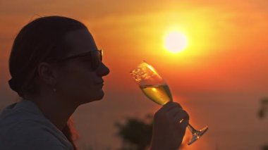 Güneş gözlüklü kadın yavaşça köpüklü şarap içerken sıcak portakal günbatımında Hilltop 'ta çimenlerin üzerinde otururken güneş cam ve gevşeme kavramının yanında hizalandı.