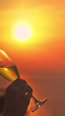 Dikey video. Güneş gözlüklü kadın yavaşça köpüklü şarap içerken sıcak portakal günbatımında Hilltop 'ta çimenlerin üzerinde otururken güneş cam ve gevşeme kavramının yanında hizalandı.