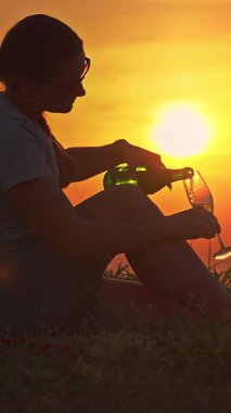 Dikey video. Çimlere Oturan Kadının Yakın Çekimi Yeşil Şişe 'den Parlak Güneş Arkaplanda, Kutlama Konsepti ile Cam' e Köpüklü Şarap Döküyor