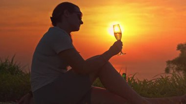 Çimenlerde oturan kadın, Güneş 'in batışına doğru elinde şampanya kadehiyle İçkinin içindeki altın yansımayı gözlemliyor.