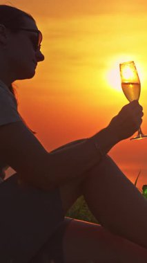 Dikey video. Çimenlerde oturan kadın, Güneş 'in batışına doğru elinde şampanya kadehiyle İçkinin içindeki altın yansımayı gözlemliyor.