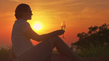 Oturan Kadın Şampanya Kadehini tutuyor ve Sıcak Altın Gün Batımına Karşı Yudumladıktan sonra Duraklıyor