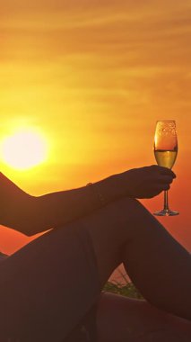 Dikey video. Oturan Kadın Şampanya Kadehini tutuyor ve Sıcak Altın Gün Batımına Karşı Yudumladıktan sonra Duraklıyor