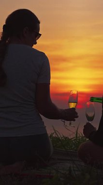 Dikey video. Tepede oturup birlikte şampanya içen çiftlerin silueti Altın Saatte Günbatımını, Romantik Anı izlerken