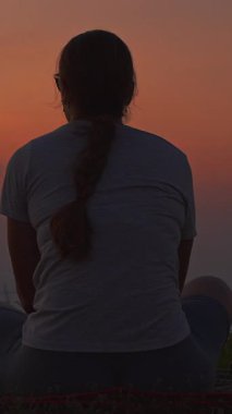 Dikey video. Piknik Mat 'te oturan ve Hilltop' tan Derin Portakal Okyanusu 'nun Günbatımını izleyen Genç Kadın' ın Arka Bakış Açısı, Yumuşak Doğal Işık ve Sakin Akşam Akşamları, Rahatlama Konsepti