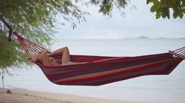 Sakin bir tropikal an yaratacak rahat bir konum bulmak için sessiz sahilin yanındaki çizgili bir hamağa inen bir kadın.