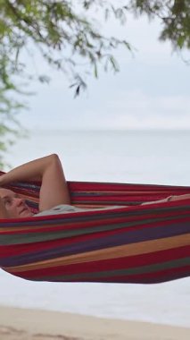 Dikey video. Sakin bir tropikal an yaratacak rahat bir konum bulmak için sessiz sahilin yanındaki çizgili bir hamağa inen bir kadın.