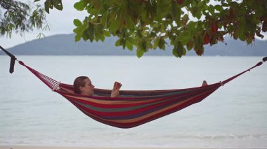 Parlak çizgili bir hamakta dinlenen bir kadın, sakin okyanus dalgalarıyla telefonuna bakarak huzurlu bir sahil anı yaratıyor.