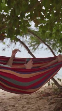 Dikey video. Bir kadın, plaj ağaçlarının altındaki hamakta uzanmış, arkasında sakin bir deniz olan, rahat bir tropikal hava yaratırken telefonuyla sosyal medyada geziniyor.