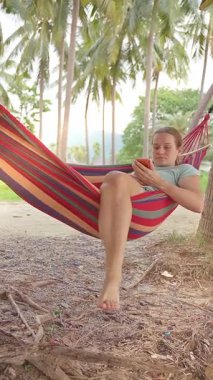 Dikey video. Palmiye ağaçları arasında çizgili bir hamakta oturan bir kadın telefonuna çıplak ayakla bakarken yere dokunuyor ve sıradan bir tropikal manzara yaratıyor.