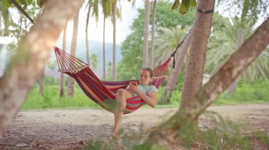 Palmiye ağaçları arasında bir hamakta dinlenen bir kadın telefonundan yeşil yaprakların arasından yumuşak bir tropikal ışık düşerken sakin bir ruh hali yaratıyor.