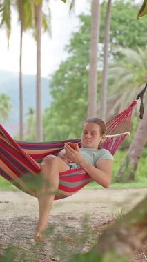 Dikey video. Palmiye ağaçları arasında bir hamakta dinlenen bir kadın telefonundan yeşil yaprakların arasından yumuşak bir tropikal ışık düşerken sakin bir ruh hali yaratıyor.