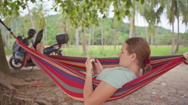 Uzun Palmiye Ağaçları ve Yumuşak Doğal Işık ile çevrili Tropik bir çayırda Smartphone 'unu gezdirirken Çizgili Hamakta Dinlenen Bir Kadın