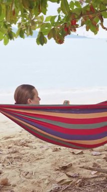 Dikey video. Sakin bir sahil seyahati atmosferi yaratan, Gölgeli Dallar Altında Smartphone 'una bakarken Hamakta Dinlenen Bir Kadın