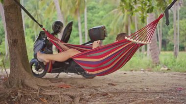 Parlak Çizgili Hamakta Uzanmış Bir Kadın Tropikal Ağaçlar Altında Bir Scooter 'ın yanında Sıcak Işıklı Rahat Bir Ruh Halinde Akıllı Telefonunu Tarayarak ve Yavaş Seyahat Hissederek