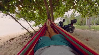 Hamak Kenarlarını Tutan Bir Adam Kum Sahili 'nin üzerinde Tropikal Ağaçlar ve Scooter' la Bir Rahatlama Kıyı Alanı Oluşturarak Rahatlıyor