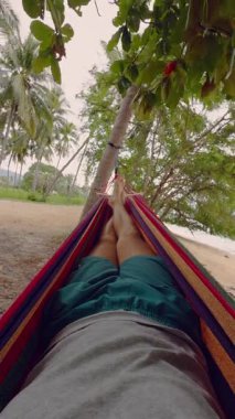 Dikey video. Sakin Dalgalar, Uzak Adalar ve Yumuşak Işık ile Denize bakan Bir Hamakta Rahatlayan Bir Adam Huzurlu Tropik Plaj Atmosferi Oluşturuyor