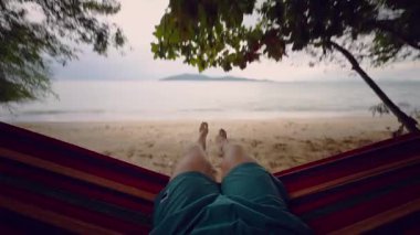 Yumuşak Işık, Sakin Dalgalar ve Tepetaklak Yapraklar ile Birinci Şahıs Manzarası 'ndan gösterilen Sessiz Tropikal bir plajda Rengarenk Hamakta Nazikçe Sallanan Bir Adam Rahatlamış Bir Kıyı Havası yaratıyor