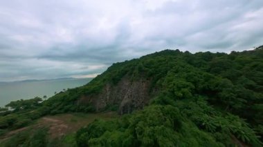 Dramatik bir FPV İHA Hız ve Macera Duyusu ile Bulutlu Gökyüzü Altında Sakin Deniz kenarında Dik Yeşil Dağ 'ı gözler önüne seriyor