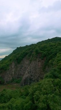 Dikey video. Dramatik bir FPV İHA Hız ve Macera Duyusu ile Bulutlu Gökyüzü Altında Sakin Deniz kenarında Dik Yeşil Dağ 'ı gözler önüne seriyor