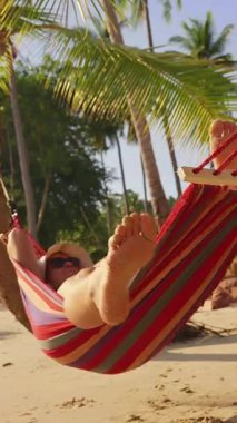 Dikey video. Hamaktaki Kadın Ayakları 'nın Düşük Açılı Görünümü Sandy Plajı' ndaki Palmiye Ağaçları Altında Tropikal Rahatlamanın Sinema Perspektifi