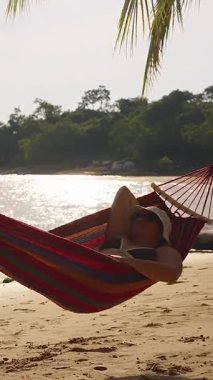 Dikey video. Hamakta yatan kadın, güneş ışığının suya yansımasıyla, huzurlu tropikal kaçış ve kıyı yaşam tarzıyla