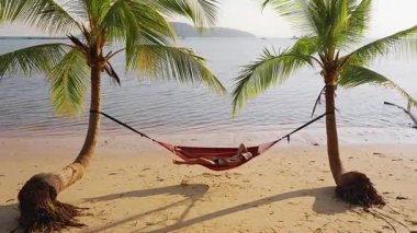 Kumsaldaki iki palmiye ağacının arasındaki renkli hamakta dinlenen sakin bir deniz ve uzun gölgeli bir tatil atmosferi yaratan bir kadın.