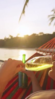 Dikey video. Güneş batarken hamakta şampanya içen bir kadın sıcak hava ve tatil havası yaratıyor.
