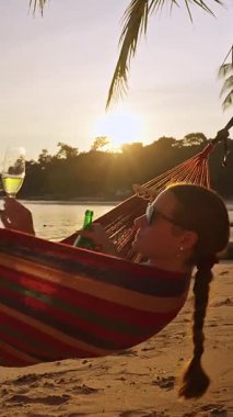 Dikey video. Şampanyasını içen kadın, güneşin batışında tropikal sahilde uzanırken altın ışık ve sakin denizle dinlendirici bir tatil havası yaratıyor.