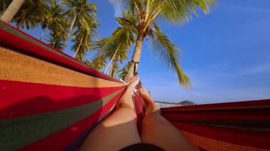 Huzurlu bir pov görüntüsü, parlak güneş ışığı ve berrak mavi gökyüzünün oluşturduğu uzun palmiye ağaçlarının altında dinlenen gevşek bacakları gösteriyor.