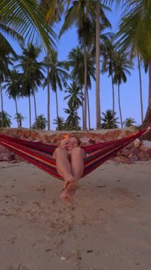 Dikey video. Bir kadın kumlu plajda bir hamakta dinleniyor. Uzun palmiye ağaçlarının altındaki telefonuna bakıyor. Yumuşak bir akşam ışığı gökyüzünü boyuyor ve huzurlu bir tropik dinlenme ortamı yaratıyor.