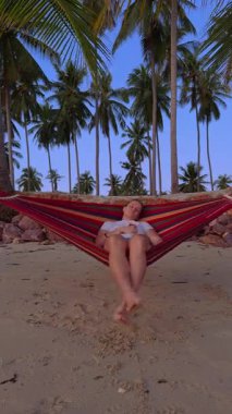 Dikey video. Bir kadın, kumlu bir sahilde, açık mavi gökyüzünün altındaki yüksek palmiye ağaçlarıyla çevrili bir hamakta uyuyor ve sakin bir tropikal rahatlama havası yaratıyor.