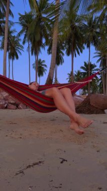 Dikey video. Bir kadın hamakta dinleniyor gözleri kapalı kumlu bir sahilde yumuşak akşam ışığı altında uzun palmiye ağaçlarının altında sakin bir tropikal dinlenme hissi yaratıyor.