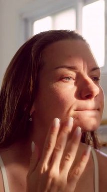 Dikey video. Aynadaki Yansımayı Yumuşak Doğal Işıkla Gözlemlerken Yüze Dokunan Kadın, Sabah Rutini, Cilt Bakımı, Özenli Özbakım ve Sakin Yaşam Tarzı