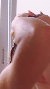 Dikey video. Yeşil Tropikal Görüş ile Pencerenin yanında duran Islak Saçlara Masaj Yapan Kadın, Doğal Işık Sakinlik Duşu Rutini Oluşturuyor