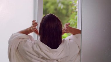 Duştan sonra havluyu takarken saçlarını düzelten kadının arka görünüşü, doğal ışık ve yeşil arka plan Rahat Sabah rutinini oluşturur