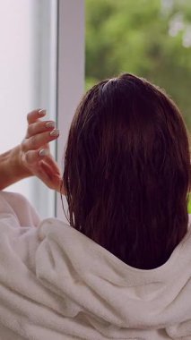 Dikey video. Duştan sonra havluyu takarken saçlarını düzelten kadının arka görünüşü, doğal ışık ve yeşil arka plan Rahat Sabah rutinini oluşturur