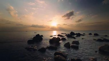 Karanlık Kıyı Kayaları ve Altın Güneş Yansıması ile Sakin Okyanus Üzerindeki Gün batımı Gökyüzünde hareket eden dramatik bulutlar Huzurlu Doğa ve Seyahat Havası yaratıyor