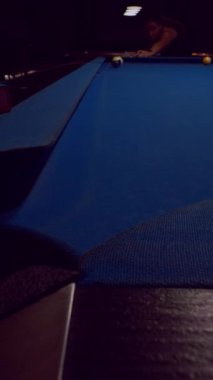 Dikey video. Geniş açılı mavi bilardo masası. Kadın karanlık kulüp iç mekanında çekim hazırlıyor. Odaklanma ve konsantrasyon gösteriyor.