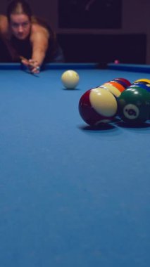 Dikey video. Cue Ball 'a nişan alan kadın mavi bilardo masasında toplar profesyonel odaklanma ve kontrol gösteriyor.