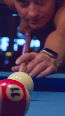 Dikey video. Kırmızı ve beyaz bilardo topuna nişan alan kadın düşük açıyla sığ derinlikten yakalandı.