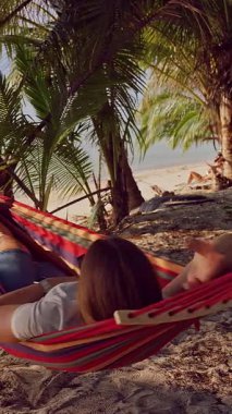 Dikey video. Hamaktan, tropik plajın yakınındaki palmiye ağaçlarının altında uzanan ilk kişi rahatlama hissi yaratıyor.