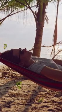 Dikey video. Hamakta deniz kenarında dinlenen bir kadın. Palmiye ağaçları ve yumuşak güneş ışığı suyu yansıtıyor. Sakin bir tropik tatil havası yaratıyor.