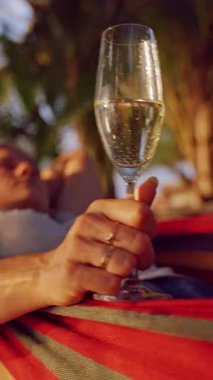 Dikey video. Tropikal palmiye yaprakları etrafındaki sakin kumlu sahili sararken hamakta uzanan bir kadın sıcak günbatımına doğru şampanya kadehini kaldırıyor.